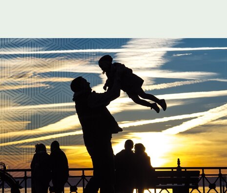 Ein Mann hebt ein Kind im Sonnenuntergang hoch. Die beiden sind nur als schwarze Silhouetten zu erkennen.