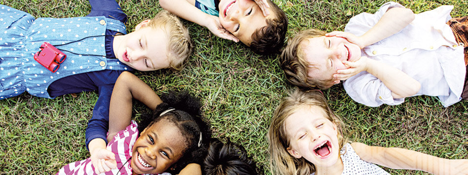 Sechs Kinder unterschiedlicher Nationalitäten liegt kreisförmig angeordnet und mit den Köpfen zueinander. Sie lachen.
