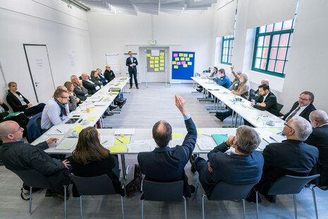 Weitwinkelaufnahme eines Konferenzraumes während eines Workshops. Ein Teilnehmer hebt die Hand.